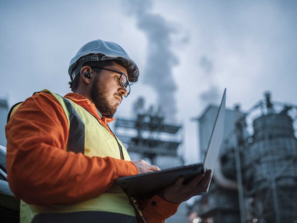 Worker using a laptop; factory in background.